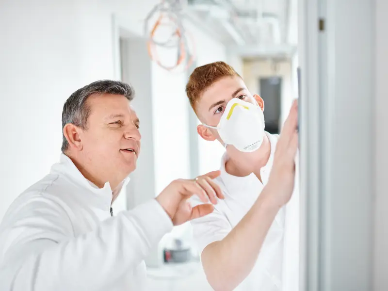 Maler zeigt einem Auszubildenden wie er etwas an der Wand machen soll
