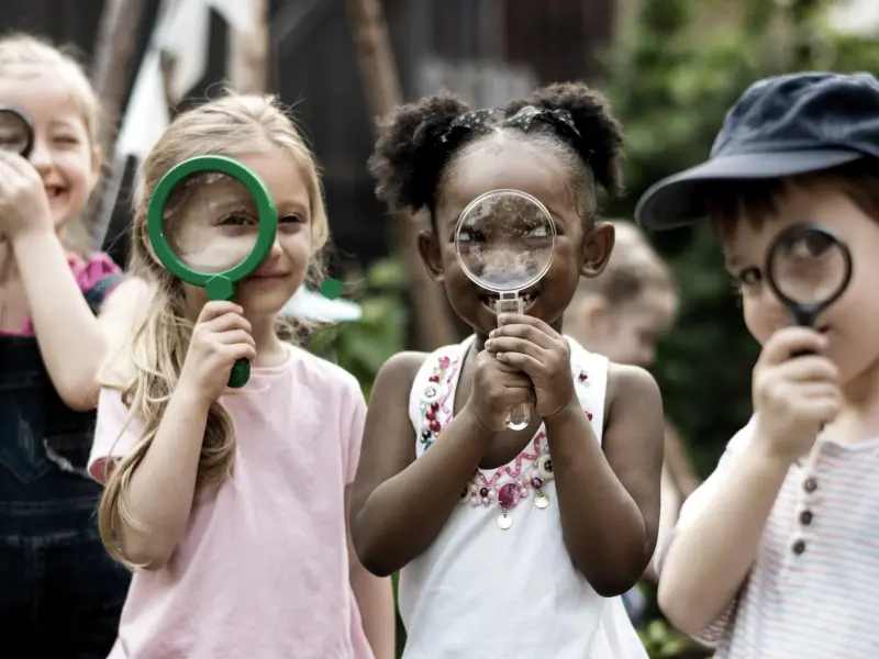 Vier verschiedene Kleinkinder schauen durch eine Lupe in die Kamera
