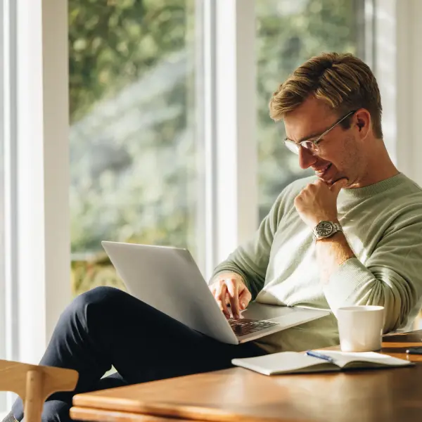  Eine Person sitzt entspannt an einem Holztisch in einem hellen Raum mit großen Fenstern, arbeitet an einem Laptop und hat eine Tasse Kaffee sowie ein Notizbuch vor sich. Im Hintergrund ist grüne Natur sichtbar.