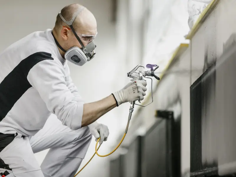 Maler mit Maske sprüht mit einem Sprühgerät Lack an einen Türrahmen