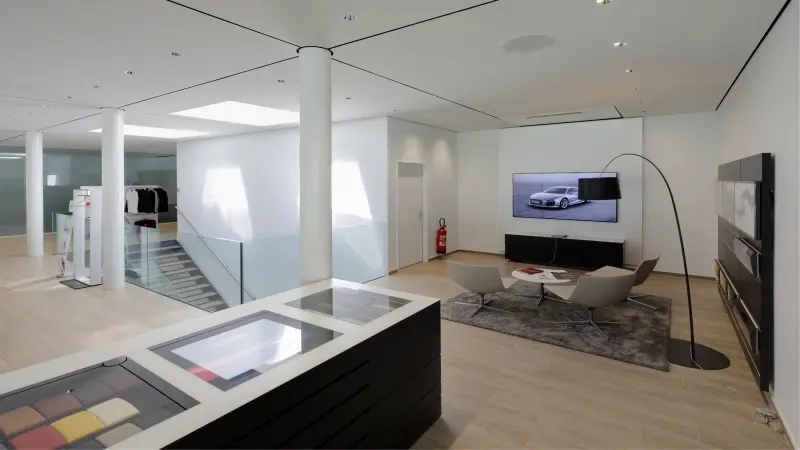 Modern eingerichteter Innenraum mit hellen Wänden, einer gemütlichen Sitzecke, einem Flachbildschirm, einer Treppe mit Glasgeländer und einer Auslage mit Materialmustern.