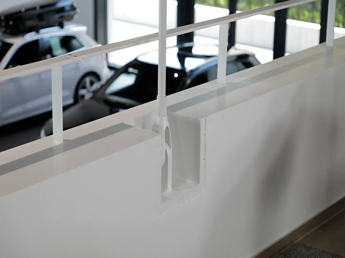 Detailaufnahme eines weißen Geländers mit klarer, moderner Verarbeitung, im Hintergrund unscharfe Fahrzeuge in einem Ausstellungsraum.