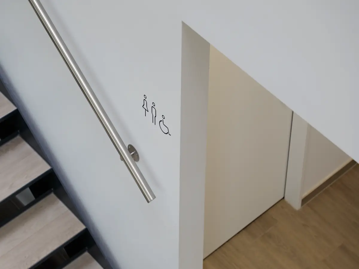 Minimalistisches Treppenhausdesign mit Holzstufen, Edelstahlhandlauf und Piktogrammen für Toiletten, einschließlich barrierefreier Zugang.