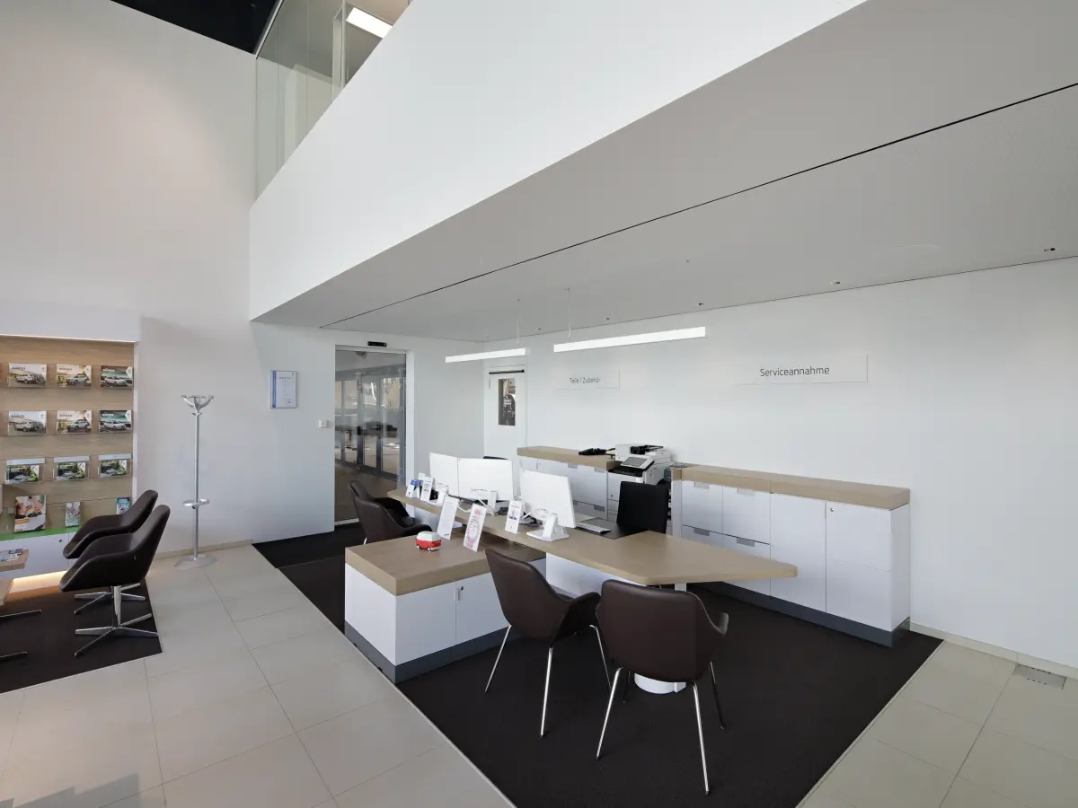 Heller, moderner Empfangsbereich mit Schreibtischen, Stühlen und Beschilderung für "Serviceannahme" sowie einer Präsentationswand mit Broschüren.