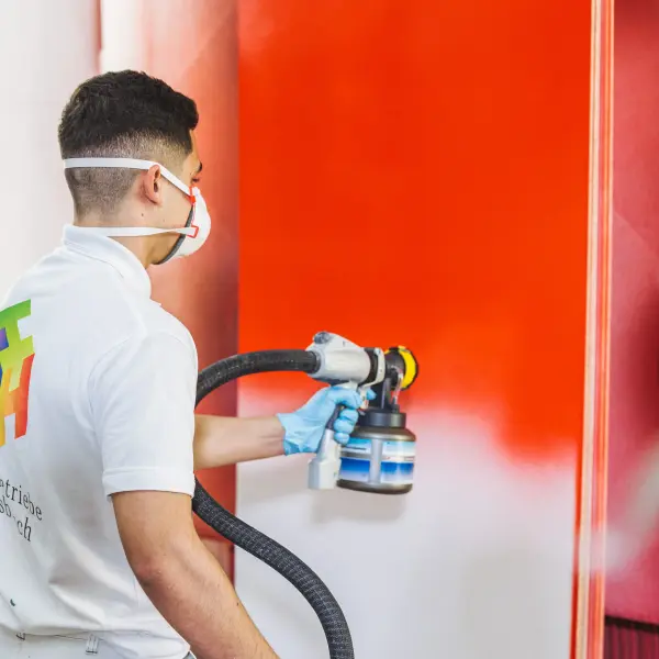 Eine Person in Arbeitskleidung und blauen Handschuhen verwendet eine Farbspritzpistole, um eine glatte Oberfläche in leuchtendem Rot zu lackieren. Im Hintergrund ist ein Teil einer Spritzkabine erkennbar.