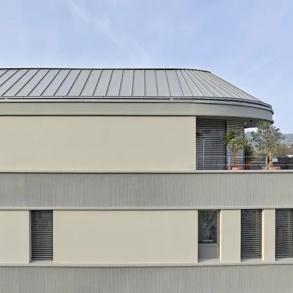  Fassade eines modernen Gebäudes mit einem geschwungenen Blechdach, hellen Putzflächen und strukturierten grauen Vertikalstreifen.