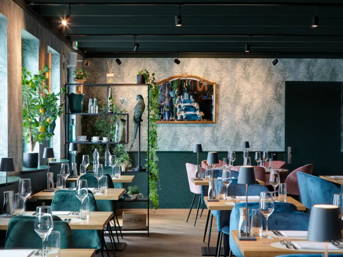 Elegantes Restaurant mit stilvollen Holztischen, grünen und rosa Samtstühlen, Palmenmuster-Wänden und dekorativen Pflanzen sowie Regalen, die ein gemütliches Ambiente schaffen.