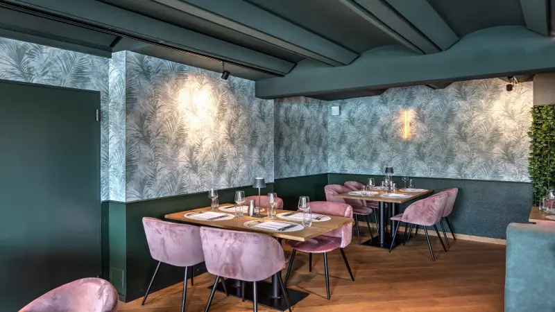 Gemütliches Restaurantinterieur mit holzfarbenen Tischen, rosa Samtstühlen und grüner Wandgestaltung mit Palmenmuster, abgerundet durch dezentes warmes Licht.