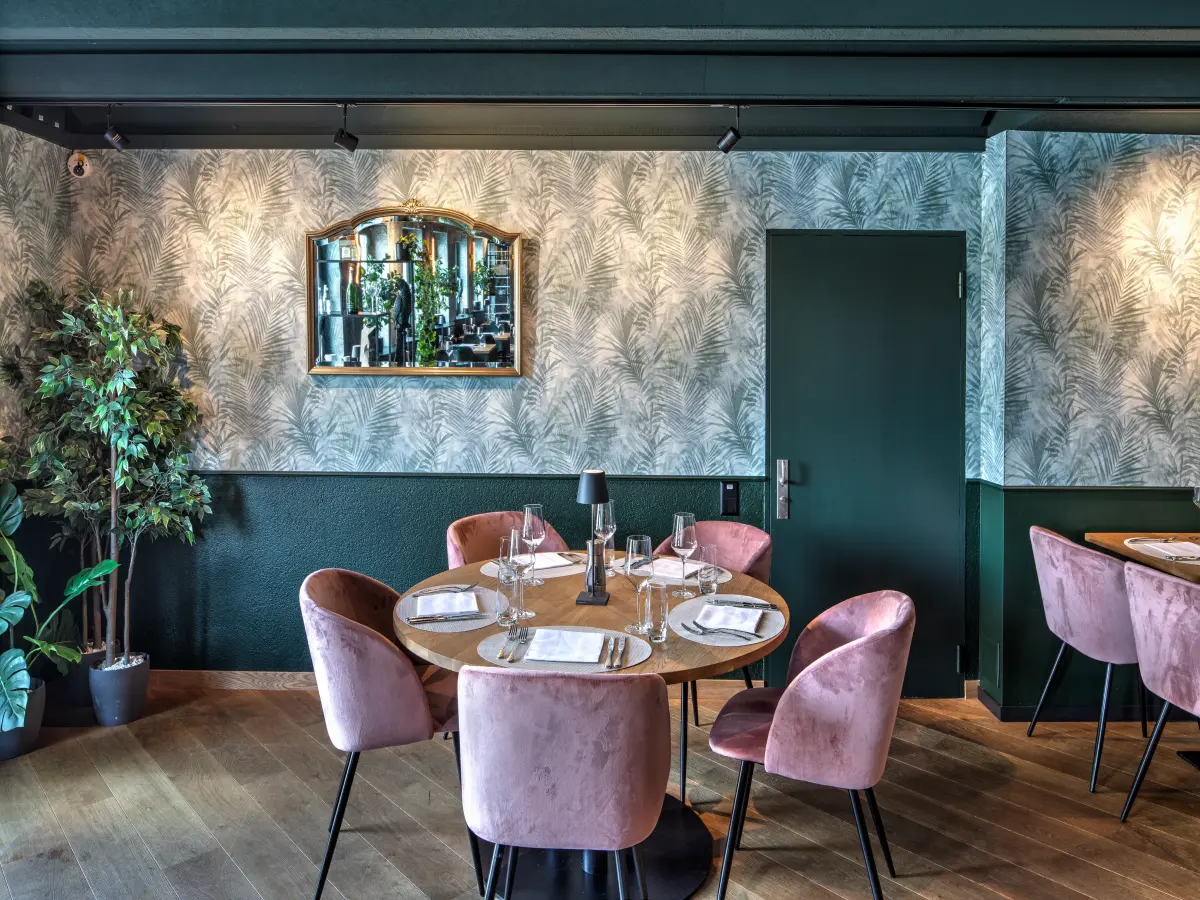 Gemütlicher Restaurantbereich mit rundem Holztisch, rosa Samtstühlen, grüner Wandgestaltung mit Palmenmuster und einem eleganten Spiegel, umgeben von dekorativen Pflanzen.