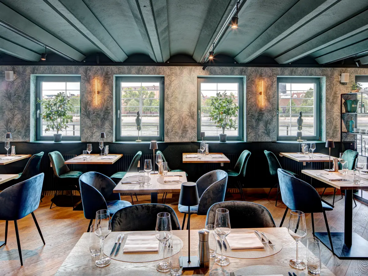 Helles Restaurant mit stilvollen Holztischen, blauen und grünen Samtstühlen, großen Fenstern mit Blick ins Freie, dekorativen Pflanzen und einer Wandgestaltung im Palmenmuster.