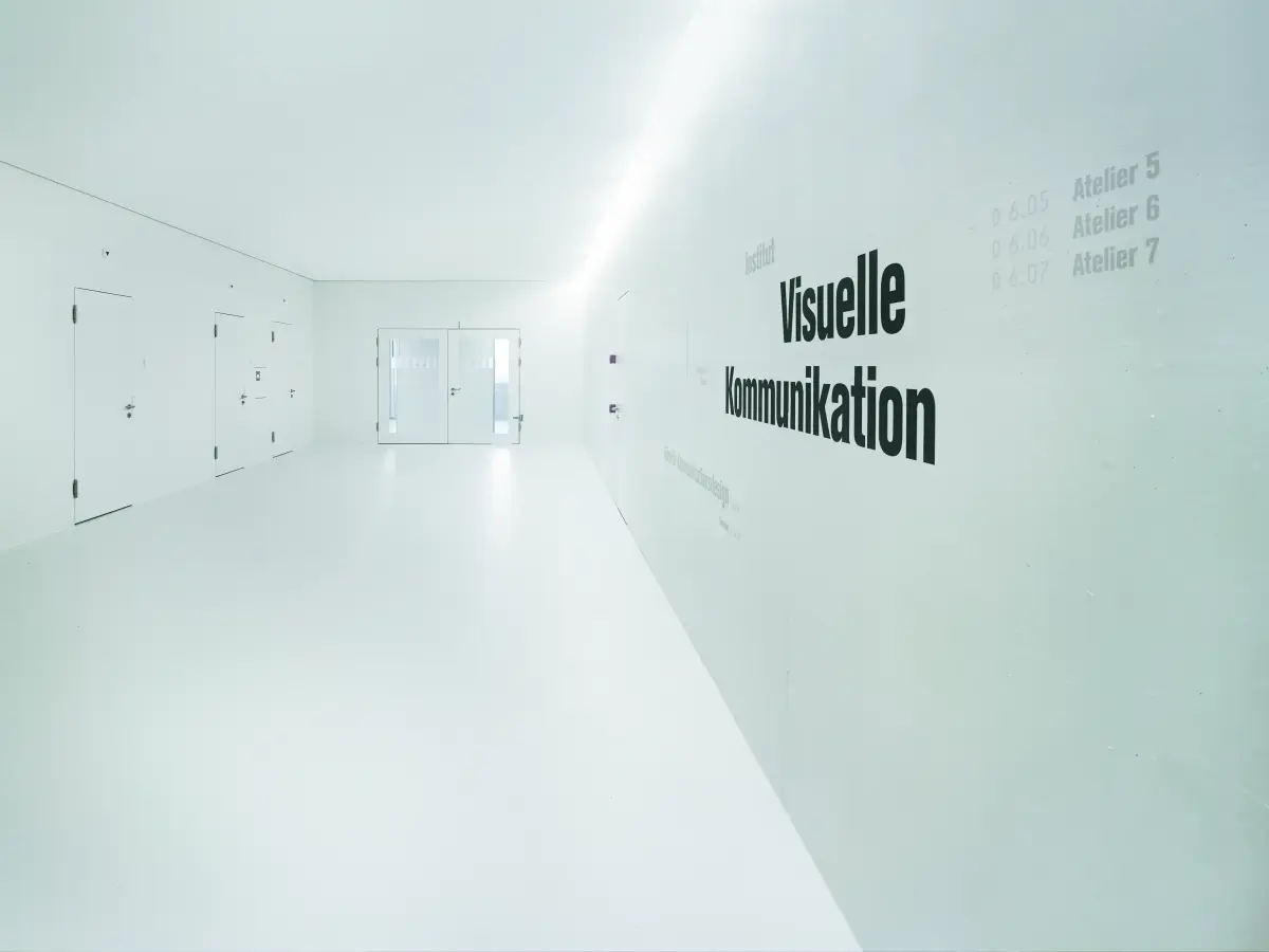 Heller, minimalistischer Flur mit weißem Boden, Türen und einer Wandaufschrift "Visuelle Kommunikation" in schwarzer Schrift.