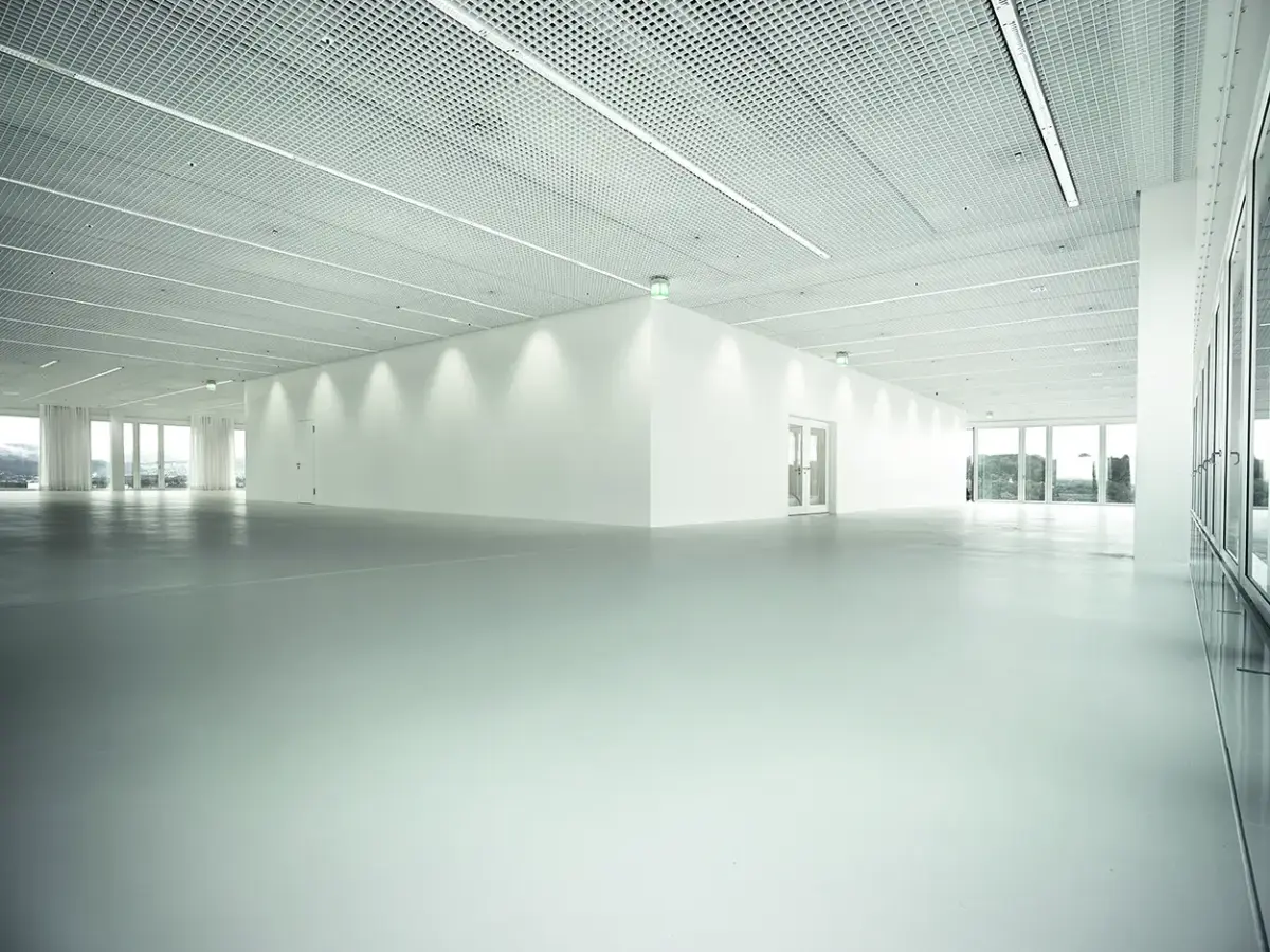 Großer, offener und heller Raum mit weißem Boden, modernen Deckenpaneelen und gläsernen Türen im Hintergrund.