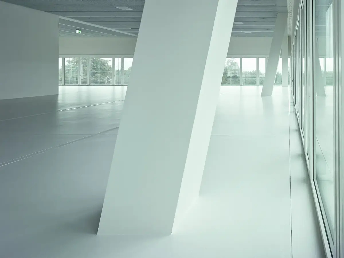 Heller, moderner Innenraum mit geneigten weißen Säulen, großen Fensterfronten und Blick ins Freie.