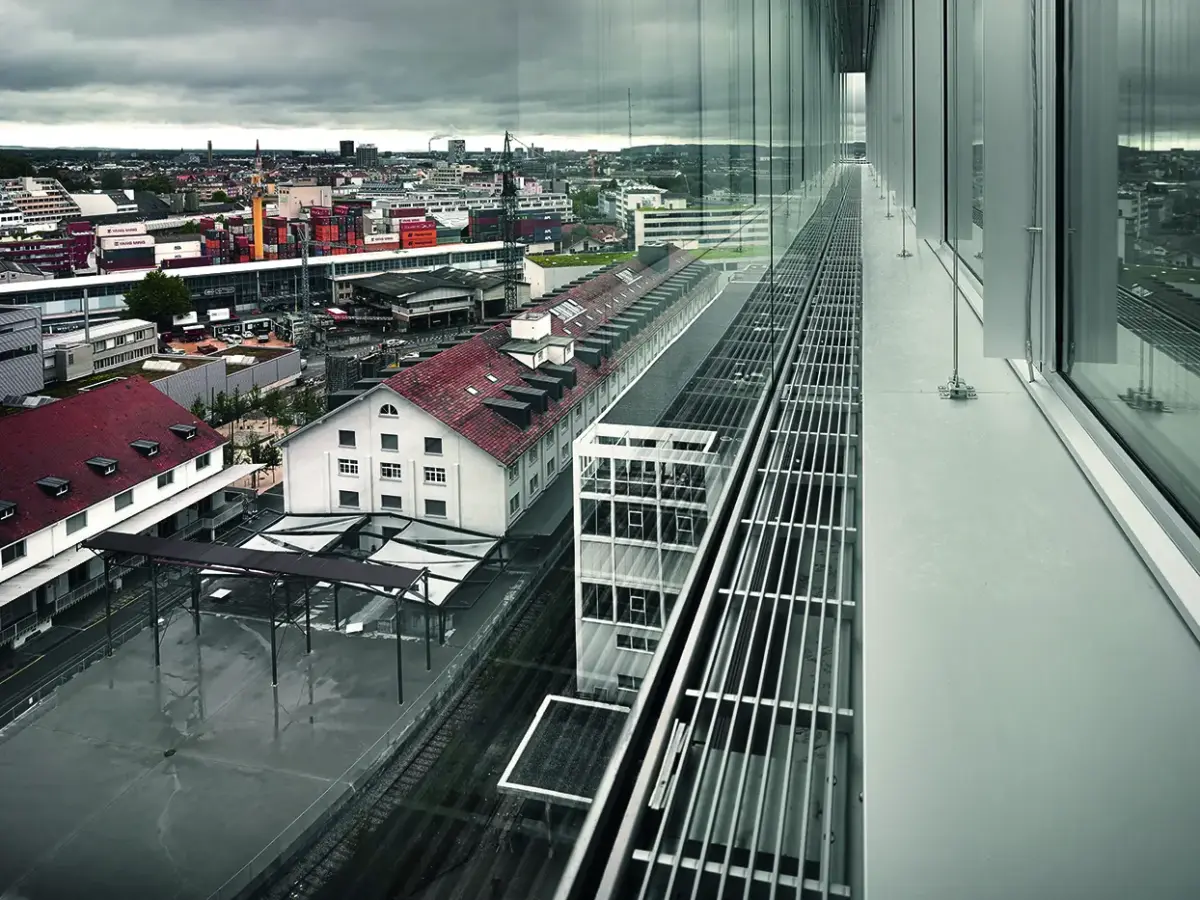 Blick von einer verglasten Gebäudefassade auf ein urbanes Industriegebiet mit roten Ziegeldächern, modernen Gebäuden und regnerischem Wetter.