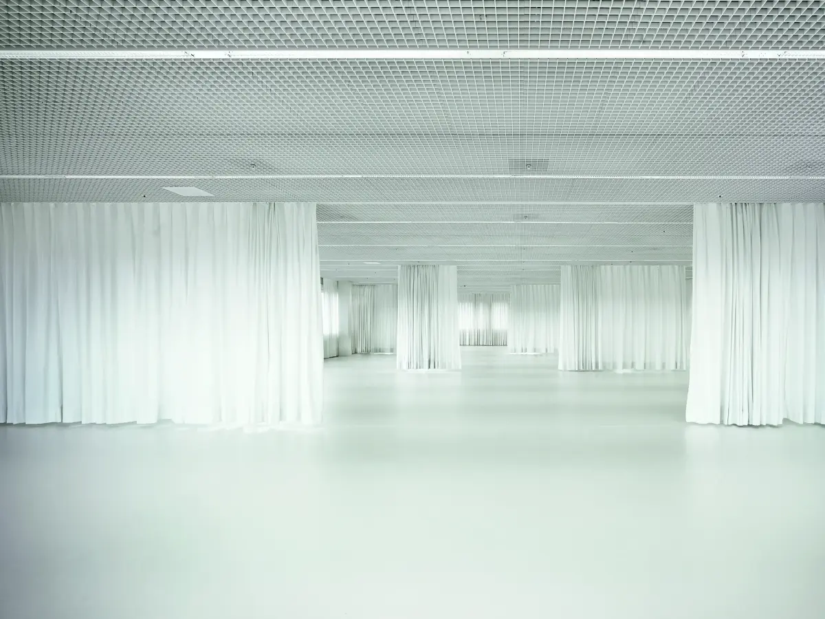 Heller, offener Raum mit weißen Vorhängen, die Bereiche abtrennen, und einer strukturierten Decke mit Gitterdesign.