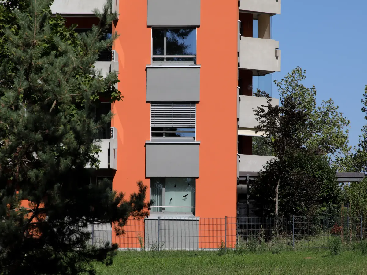 Fassadendetail eines orangefarbenen Hochhauses mit grauen Fensterläden, umgeben von Bäumen und Grünfläche.