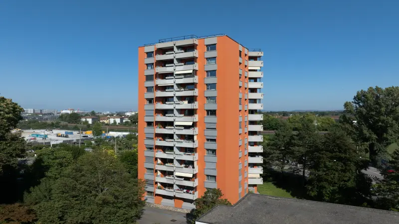 Ein orangefarbenes Hochhaus mit mehreren Balkonen auf einer Seite, umgeben von Bäumen, bei klarem blauem Himmel. Im Hintergrund sind weitere Gebäude und eine Stadtlandschaft zu sehen.