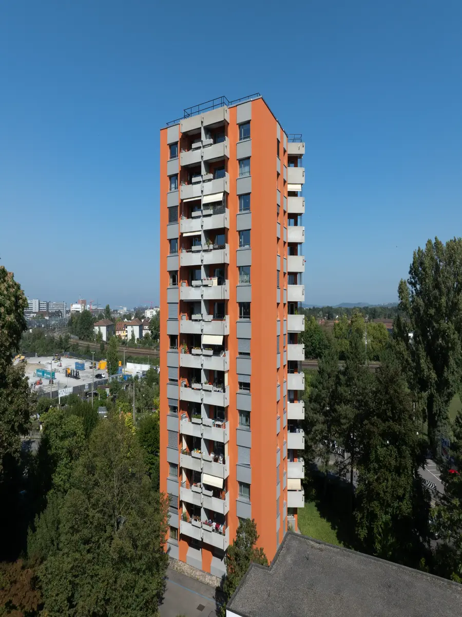 Alt-Text: Ein orangefarbenes Hochhaus mit mehreren Balkonen auf einer Seite, umgeben von Bäumen, bei klarem blauem Himmel. Im Hintergrund sind weitere Gebäude und eine Stadtlandschaft zu sehen.