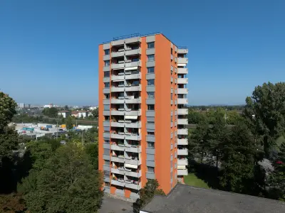 Ein orangefarbenes Hochhaus mit mehreren Balkonen auf einer Seite, umgeben von Bäumen, bei klarem blauem Himmel. Im Hintergrund sind weitere Gebäude und eine Stadtlandschaft zu sehen.