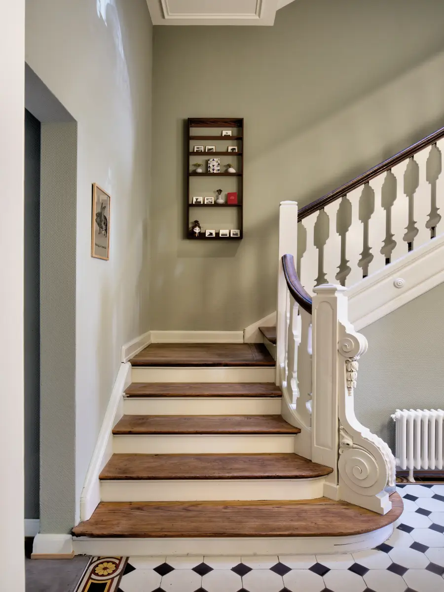 Historisches Treppenhaus mit eleganter Holztreppe, weißem Geländer und Wandregal. Der Boden besteht aus zeitlosen schwarz-weißen Fliesen.