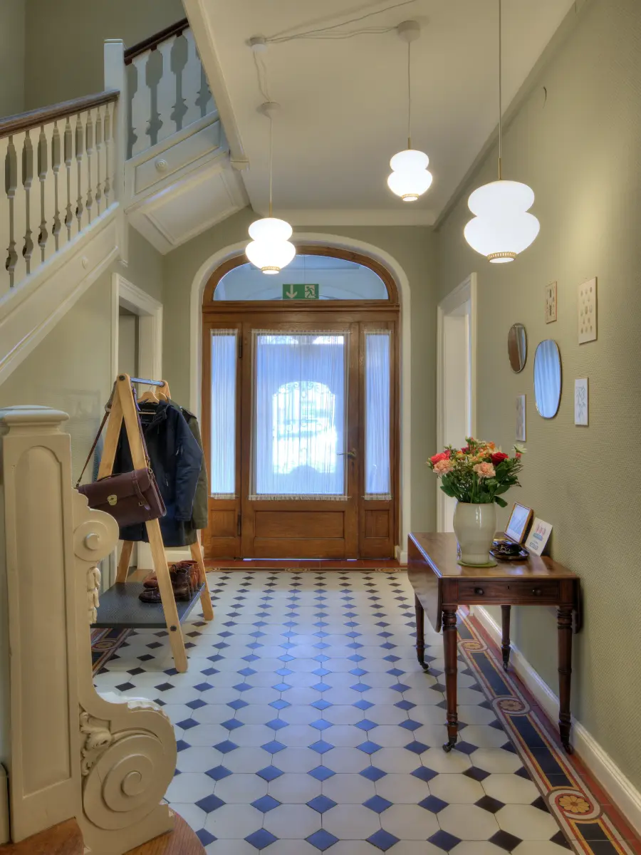 Eleganter Eingangsbereich mit klassischem schwarz-weißem Fliesenboden, einem Holzsekretär, dekorativen Lampen und einer historischen Holztreppe. Holztüren mit Glaselementen betonen den charmanten Stil.