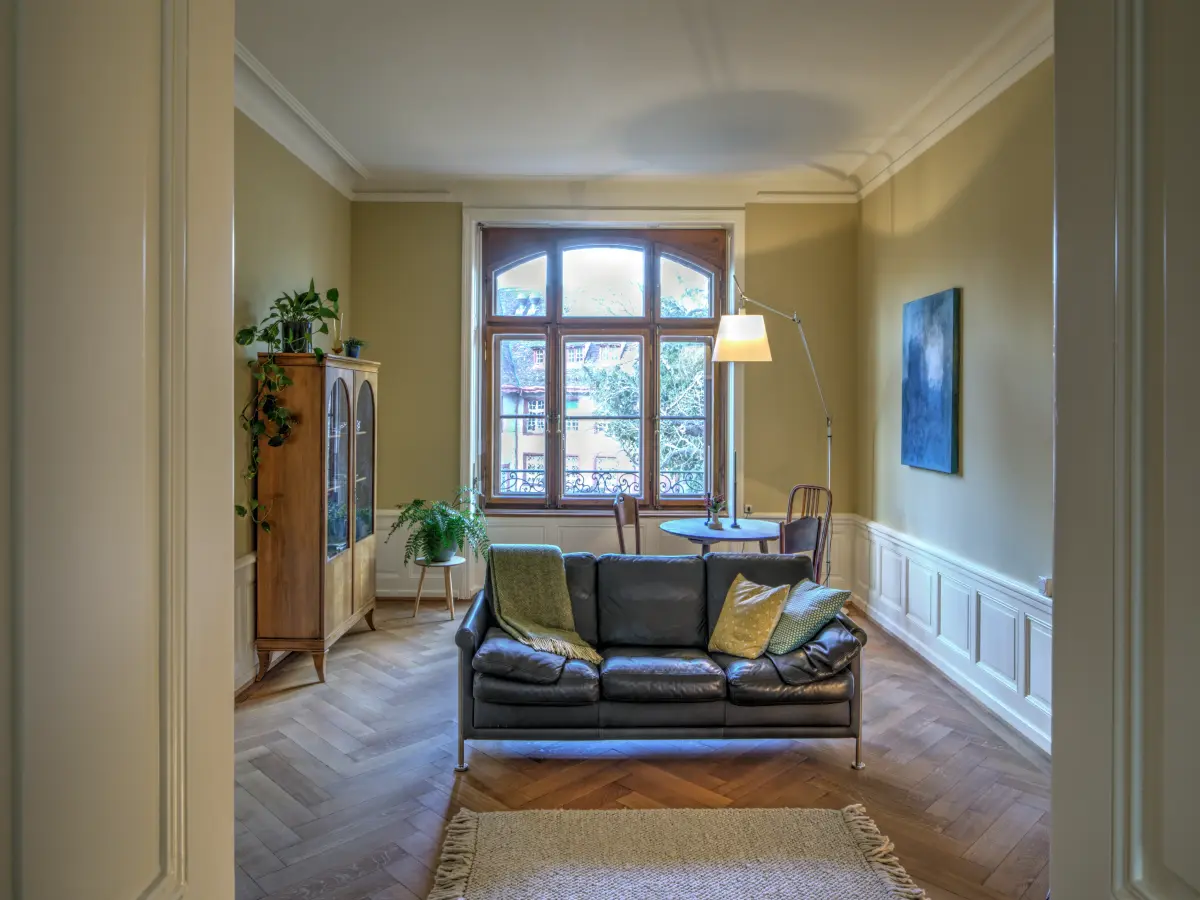 Elegantes Wohnzimmer mit großer Fensterfront, Ledersofa, Fischgrätparkett und klassischen Wandleisten. Dezente Grüntöne und Pflanzen schaffen eine gemütliche, natürliche Atmosphäre.