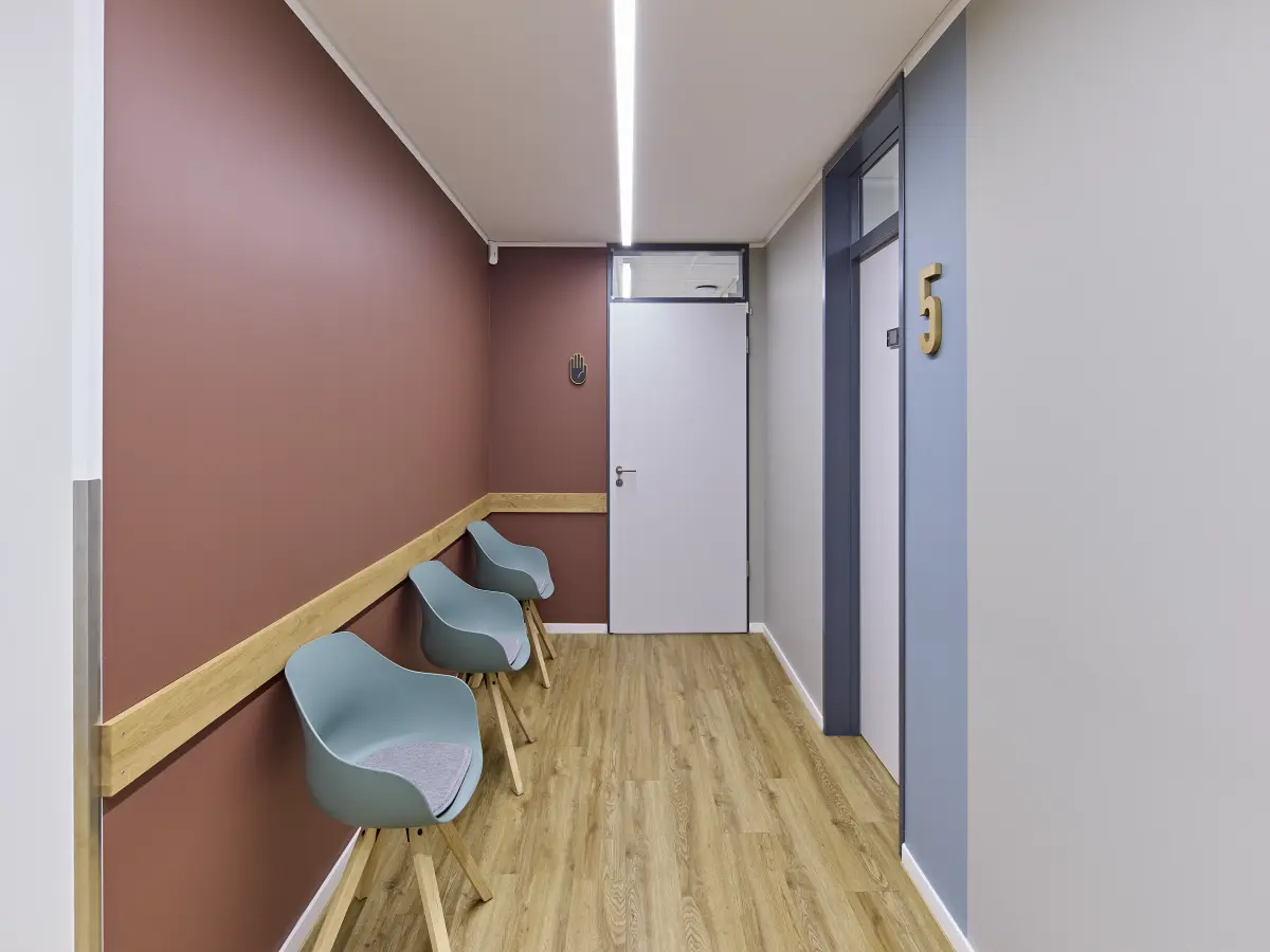 Kleiner Wartebereich mit drei Stühlen entlang einer Wand und Tür mit Raumnummer.