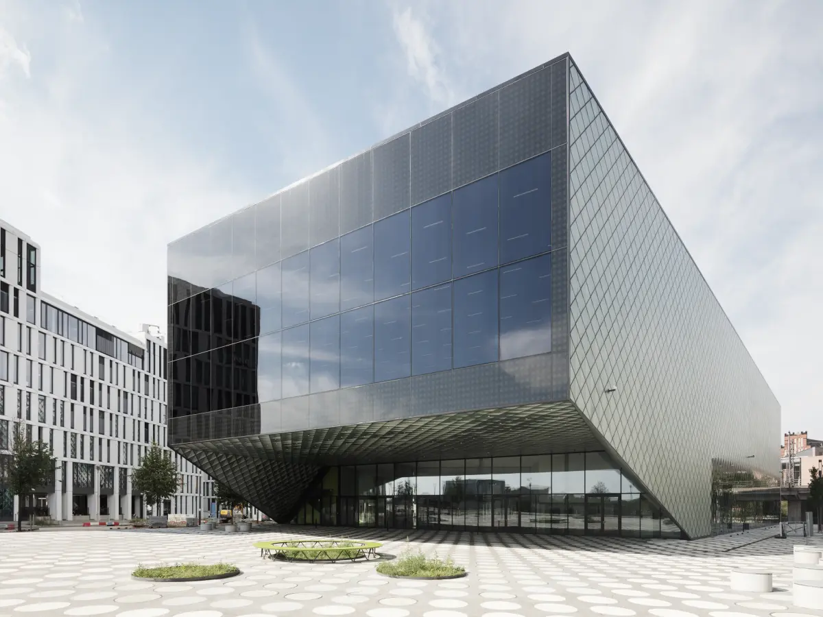 Kubischer Neubau mit dunkler Glasfassade, schräger, überdachter Eingangspartie und gepflastertem Vorplatz mit Sitzinseln.