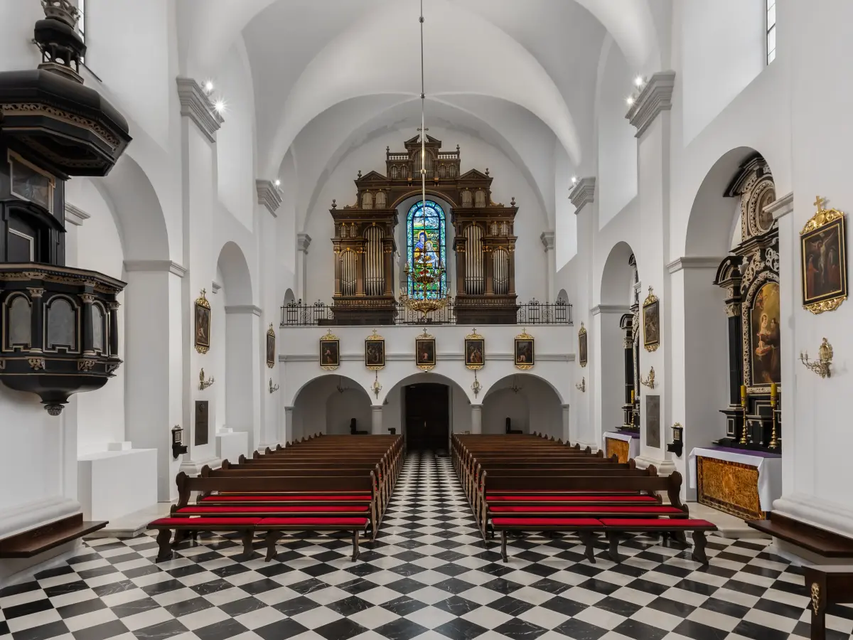 Eine große Kirche mit einem schwarz-weiß karierten Boden und Holzbänken, die zu einem verzierten Altar und einer Orgel am Ende des Ganges führen.