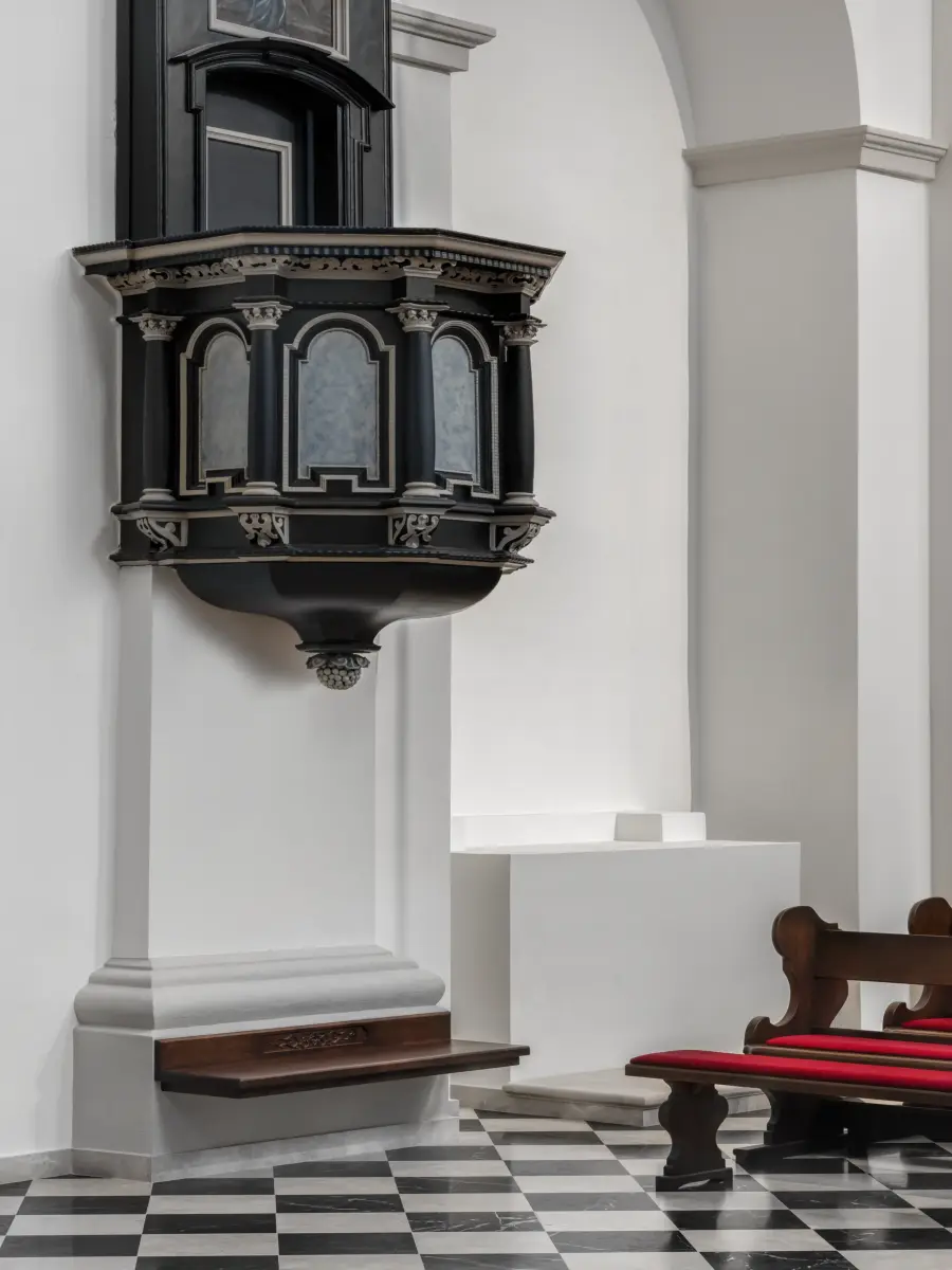 Eine schwarze, verzierte Kanzel an einer weißen Kirchenwand über einem schwarz-weiß gefliesten Boden.