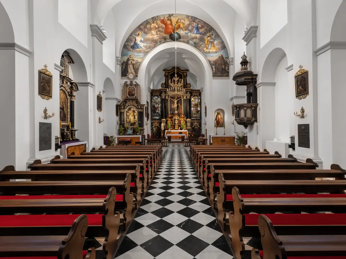 Eine große, weiße Kirche mit einem schwarz-weiß karierten Boden, Holzbänken mit roten Kissen und einem großen, verzierten Altar.