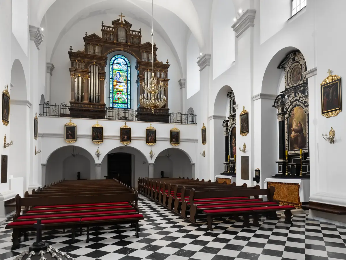 Ein barockes Kirchenschiff mit Reihen von hölzernen Kirchenbänken mit roten Polstern. Der Boden hat ein Schachbrettmuster in Schwarz-Weiß. Im Hintergrund steht eine große, reich verzierte Orgel unter einem Buntglasfenster.