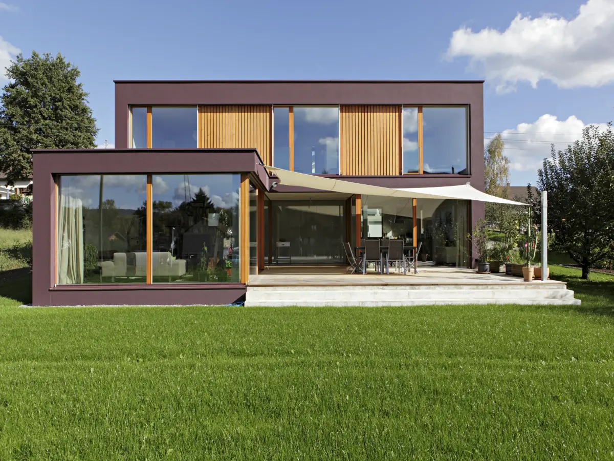 Modernes zweistöckiges Einfamilienhaus mit dunkler Bordeaux Fassade, großen Fensterfronten und hölzernen Akzenten. Eine weitläufige Terrasse mit Sonnensegel grenzt an einen gepflegten grünen Rasen.