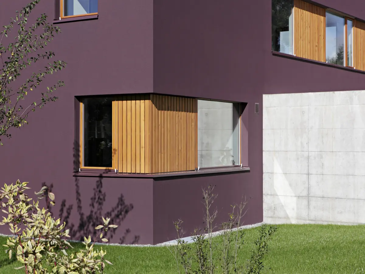 Ecke eines modernen Hauses mit lila Putzfassade. Große Fenster mit hölzernen Vertikal-Lamellen setzen Akzente, begleitet von einer Betonwand und einem gepflegten grünen Garten im Vordergrund.