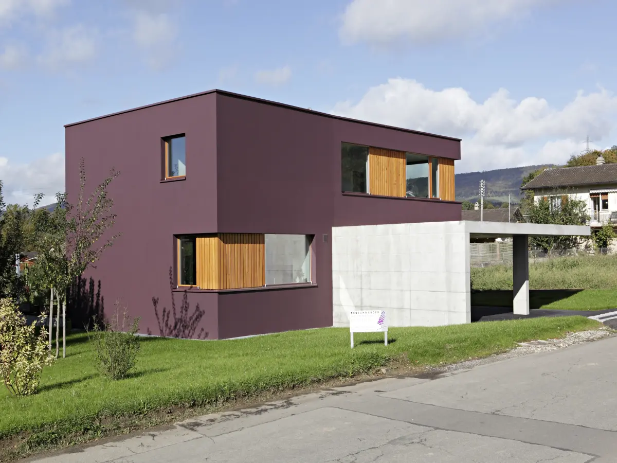 Modernes zweistöckiges Haus mit dunkel bordeauxfarbener Fassade, großen Fenstern und hölzernen Akzenten. Beton-Elemente und ein Carport fügen sich harmonisch ins Design ein. Umgeben von gepflegtem Rasen, Bäumen und einer ruhigen Nachbarschaft.