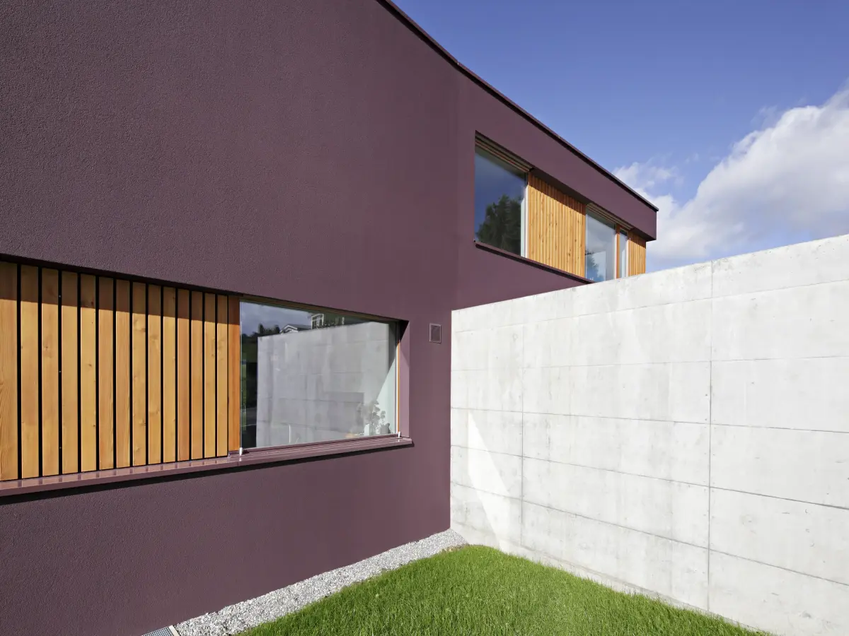 Seitenansicht eines modernen Hauses mit dunkel bordeauxfarbener Fassade. Große Fenster mit hölzernen Vertikallamellen und eine angrenzende Betonwand betonen das minimalistische Design.