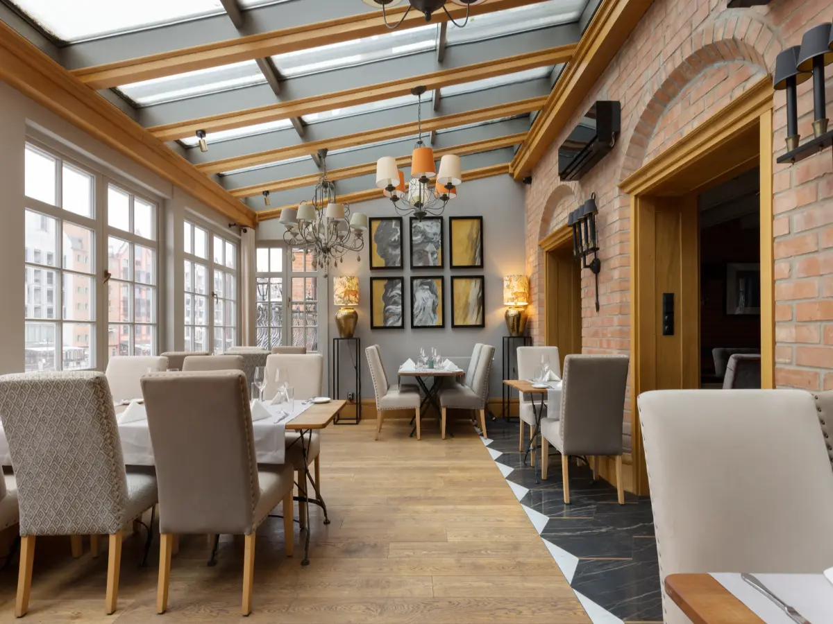 Jasne wnętrze restauracji z nakrytymi stołami, dużymi oknami i stylowymi obrazami na ścianie.