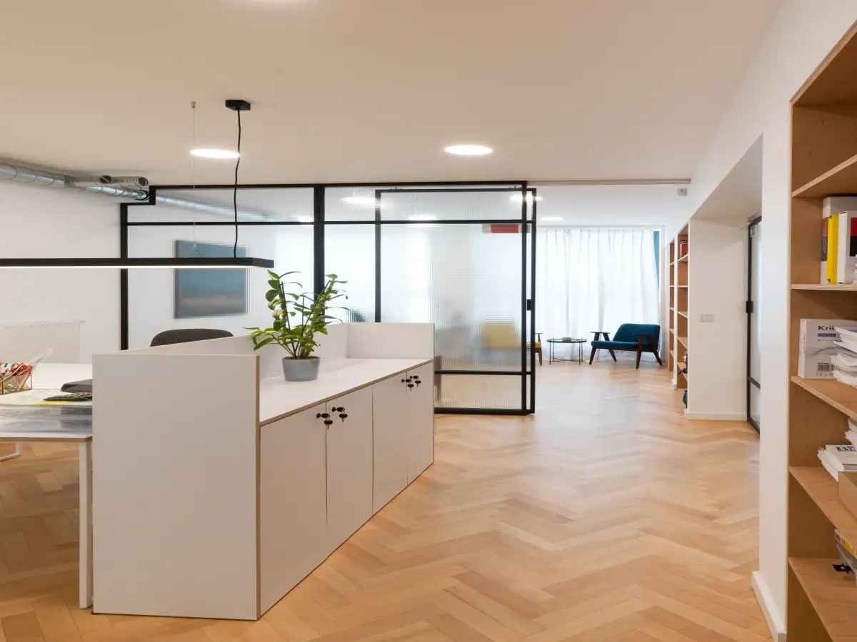 Jasna przestrzeń biurowa z drewnianym parkietem, białą wyspą recepcyjną, przeszklonymi ścianami i regałami.