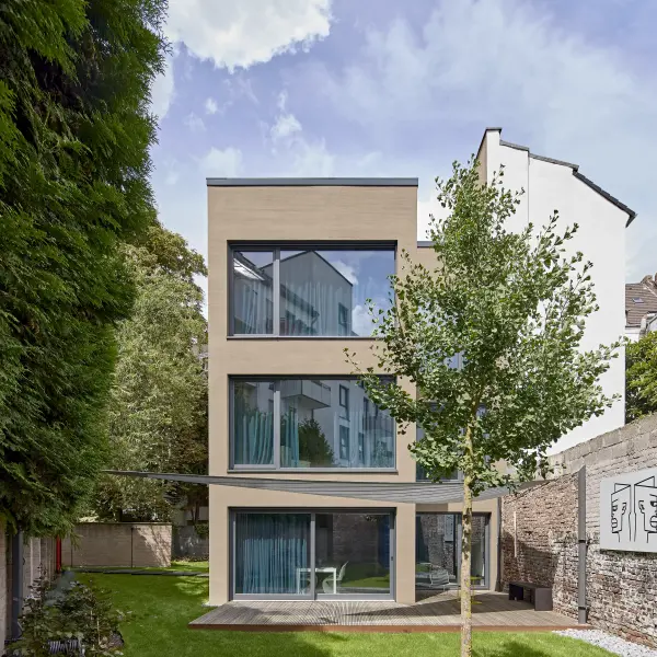  Ein modernes, dreistöckiges Wohnhaus mit beiger Fassade und großen Fensterfronten, umgeben von einem grünen Garten und einer alten Backsteinmauer.