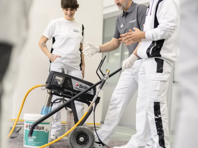  Drei Personen in Arbeitskleidung stehen um ein professionelles Farbspritzgerät von Brillux, während eine Person etwas erklärt. Im Vordergrund ist ein Farbeimer sichtbar, und der Raum hat eine helle, industrielle Atmosphäre.