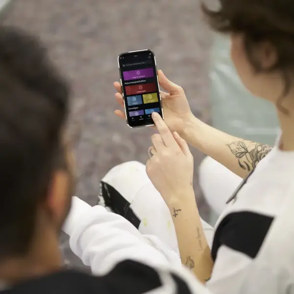 Zwei Personen in Arbeitskleidung betrachten ein Smartphone, auf dessen Bildschirm eine bunte Benutzeroberfläche mit verschiedenen Optionen zu sehen ist. Eine Person zeigt mit dem Finger auf das Display.