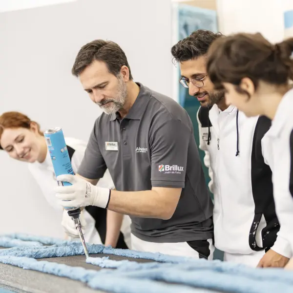 Eine Person in einem grauen Poloshirt mit Brillux-Logo demonstriert die Anwendung einer Kartuschenpistole auf einer blauen Oberfläche, während mehrere Personen in Arbeitskleidung aufmerksam zuschauen. Der Raum hat eine helle, professionelle Atmosphäre.