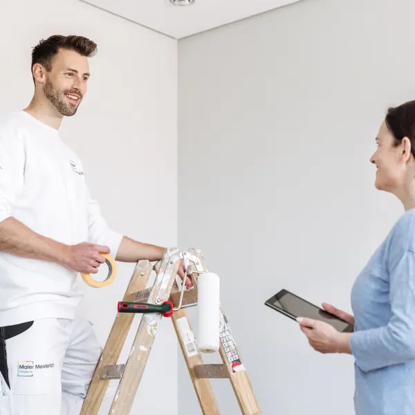 Lorem IpsuEine Person in weißer Arbeitskleidung steht auf einer Leiter und hält Malerkrepp, während eine andere Person mit einem Tablet in der Hand daneben steht. Der Raum hat helle Wände und eine minimalistische Einrichtung. Auf der Leiter befinden sich Malerutensilien wie eine Farbrolle.m