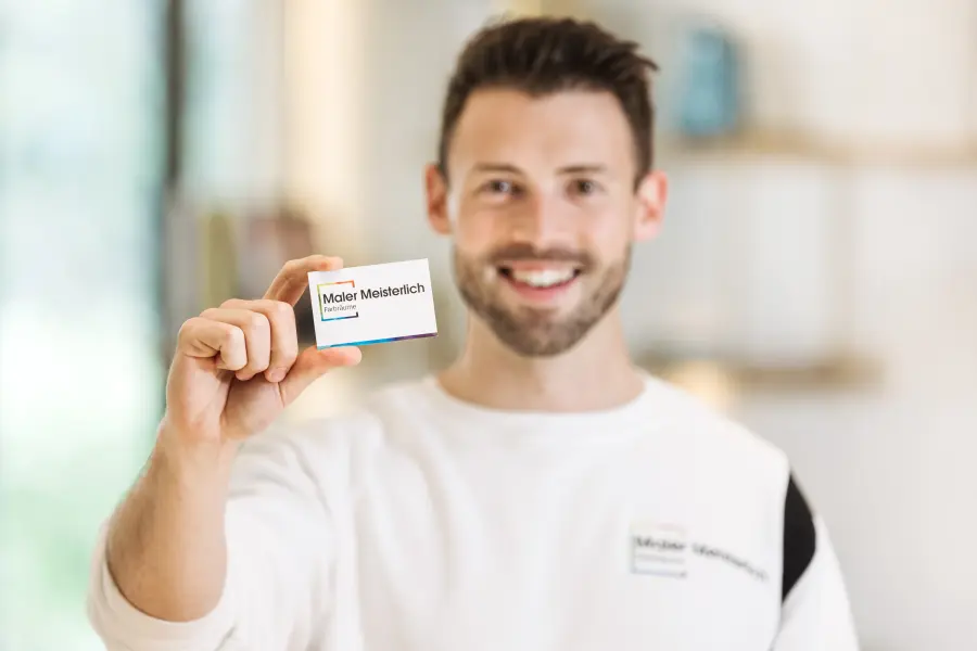 Eine Person hält eine Visitenkarte mit der Aufschrift „Maler Meisterlich“ in die Kamera. Im Hintergrund ist ein heller, unscharfer Raum mit Regalen und Dekorationen zu sehen.
