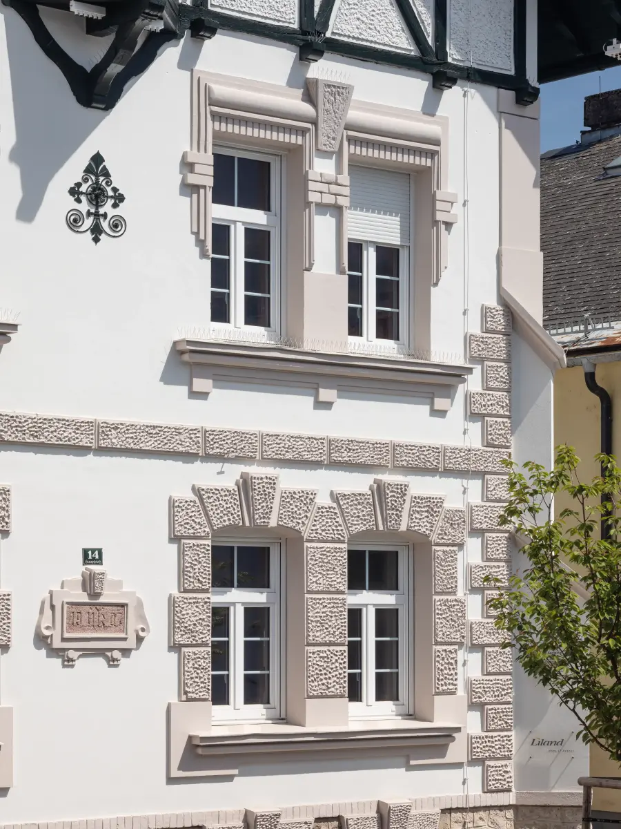 Nahaufnahme der hellen Fassade eines Hauses mit vier Fenstern und dekorativen Steinelementen.