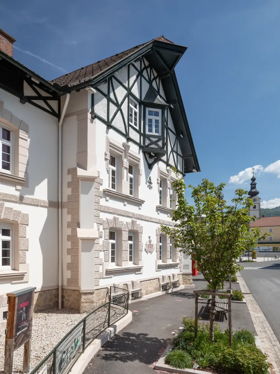  Detailansicht der hellen Fassade eines Hauses mit Fachwerk-Giebel und einem Baum davor, mit einem Kirchturm im Hintergrund.