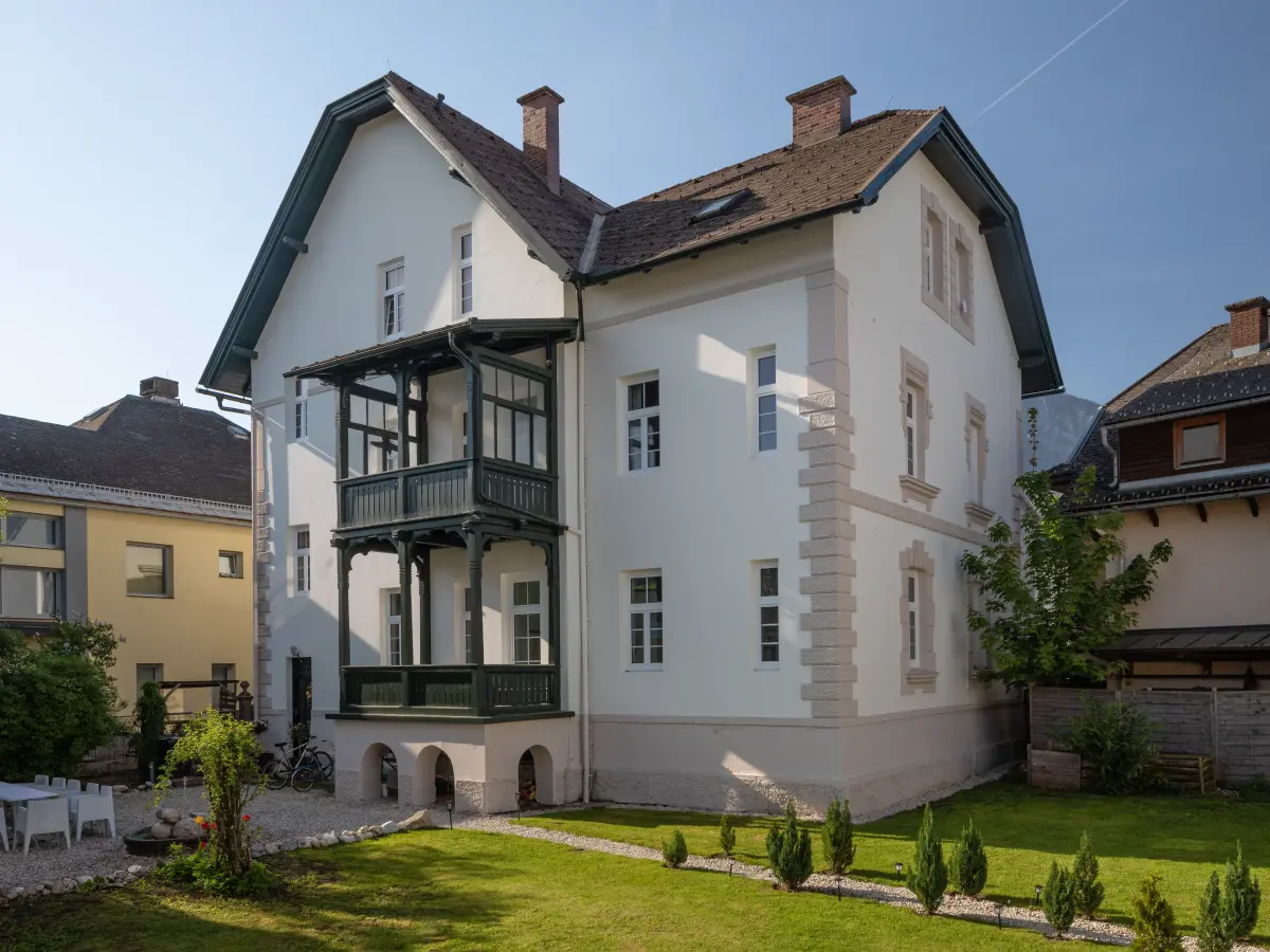  Die Rückansicht des hellen Hauses mit einem zweistöckigen, dunkelgrünen Balkon und einem gepflegten Garten.