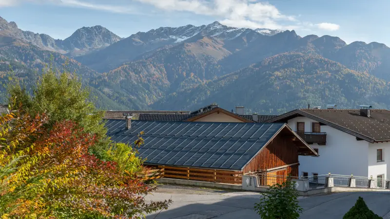 Häuser mit dunklen Dächern und Holzbalkonen vor einem Hintergrund aus herbstlich bewaldeten Alpenbergen mit schneebedeckten Gipfeln.