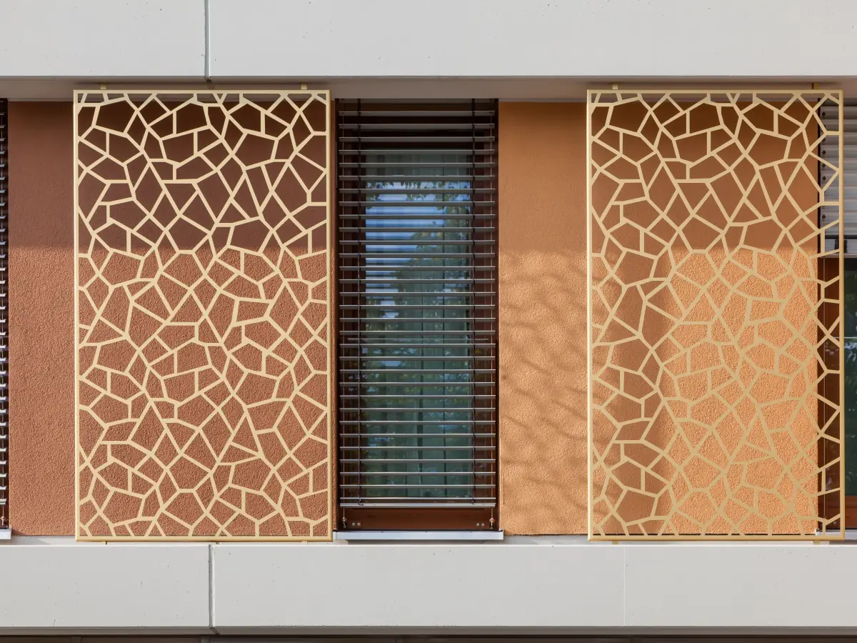 Dettaglio di facciata con schermi metallici ornamentali, finestra centrale con veneziana esterna e superficie intonacata in tonalità calde.