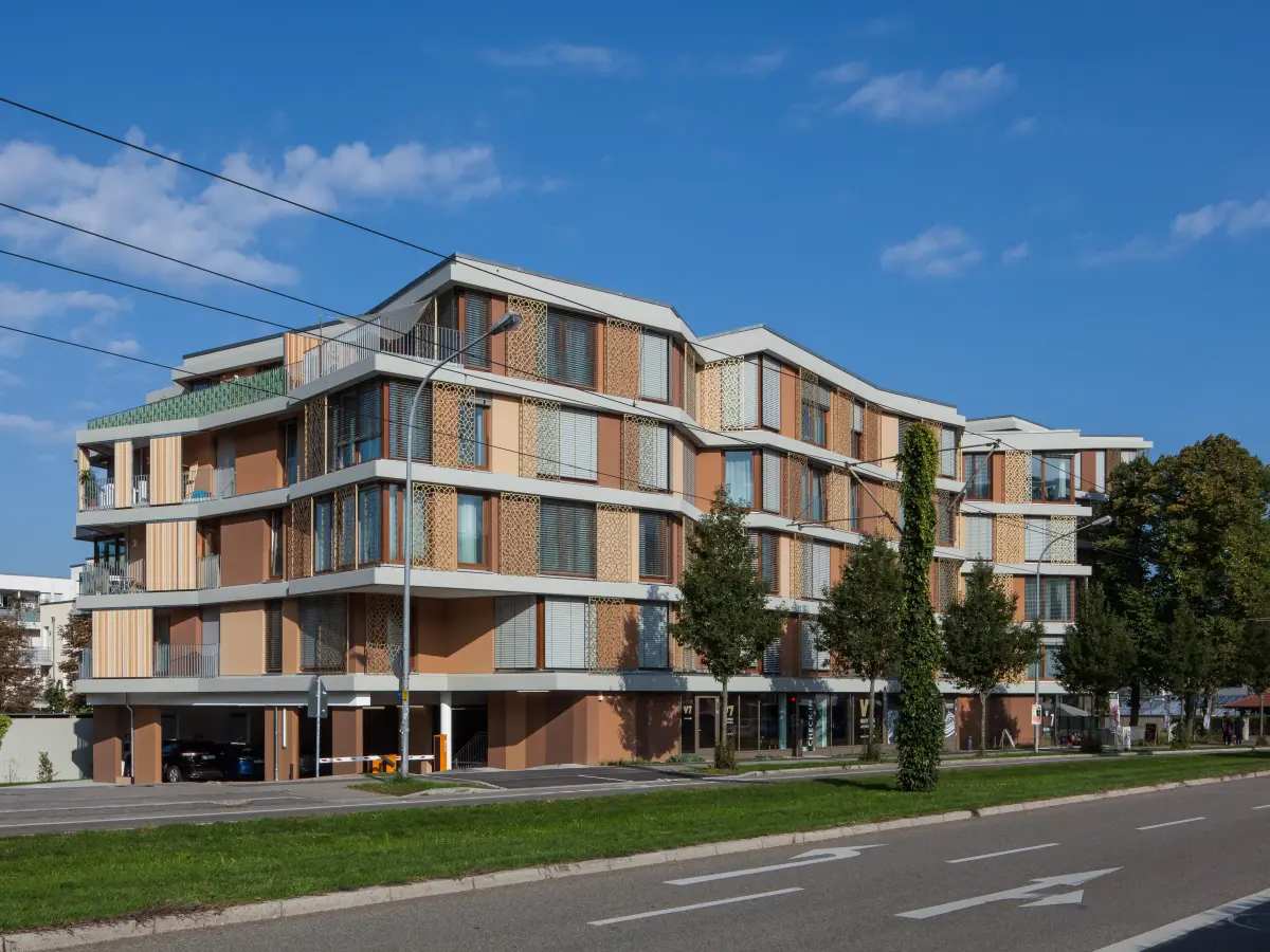 Complesso residenziale con linea del tetto curva, facciata articolata e zona al piano terra su un'ampia strada.